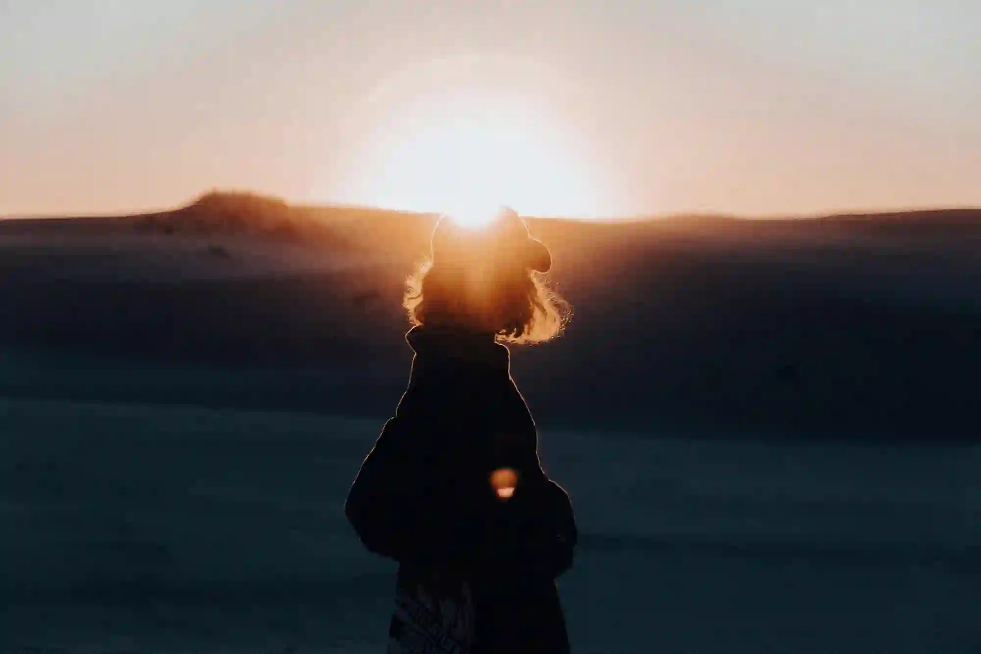 Silhouette of a person facing a sunrise over desert dunes, the sun forming a halo behind their head.